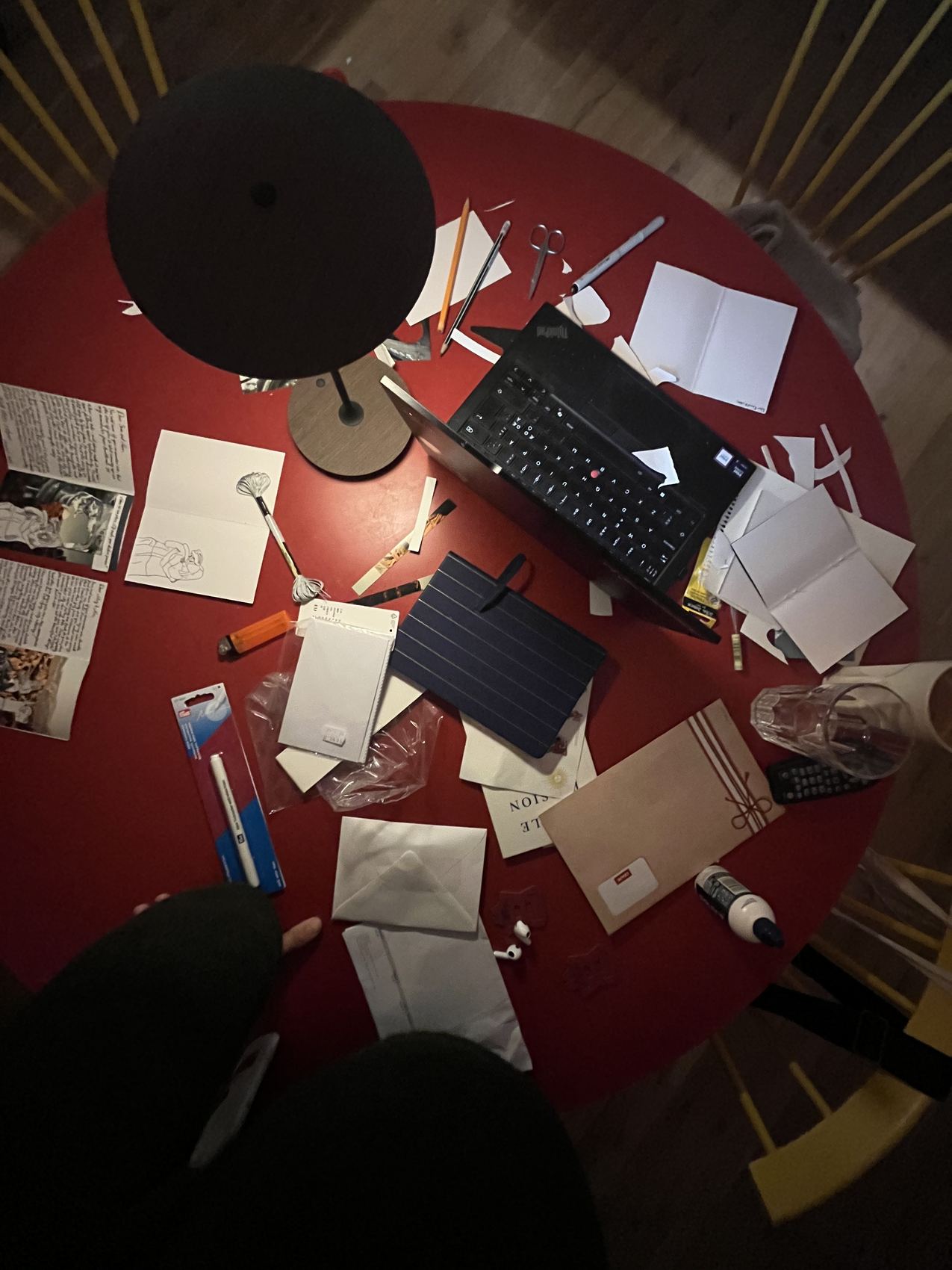 The red dinner table mid-project: cut paper, pens, scissors and part-made cards spread around a laptop.